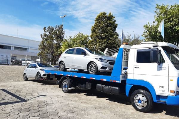 Carro branco sobre plataforma de guincho azul e branco em área aberta.